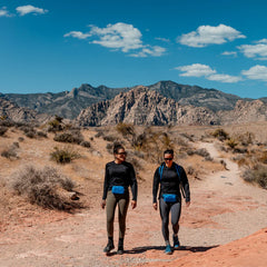 Two women hiking in desert terrain with mountains and blue sky, wearing athletic gear and blue waist packs