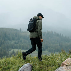 Clad in dark apparel with the Merino Challenge Socks featuring a protective Trail Cuff by GORUCK, an individual strides across a grassy hilltop enveloped by misty, forested mountains. The overcast sky enhances the serene and secluded ambiance.