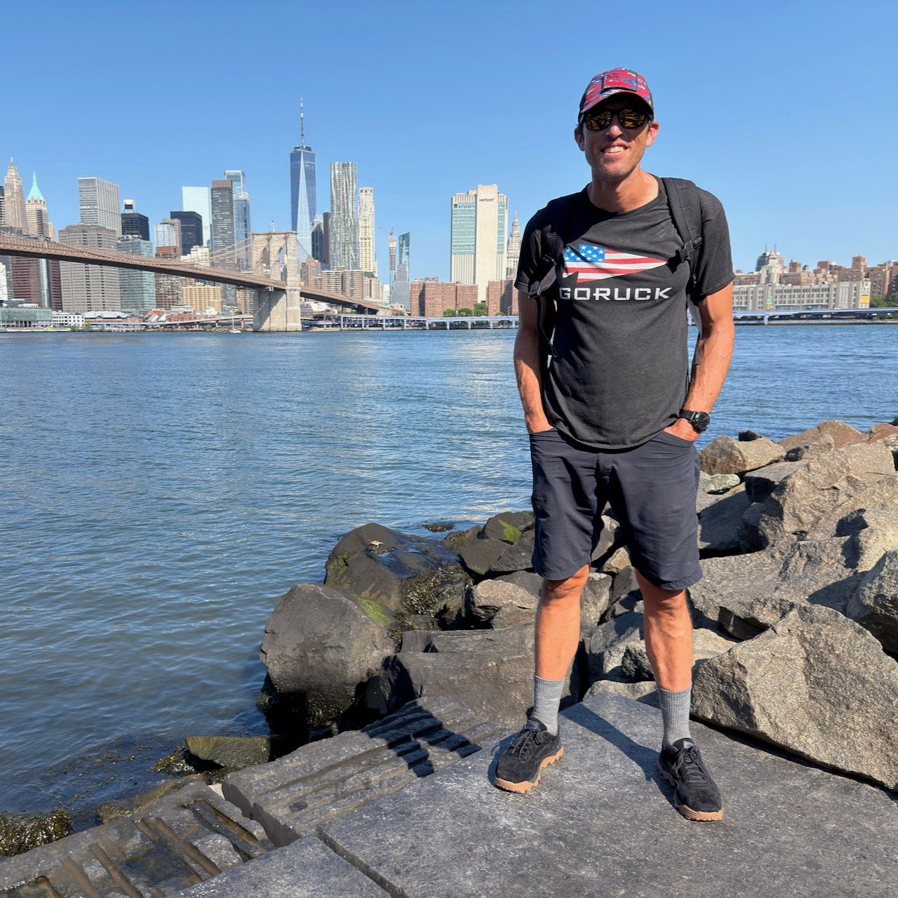 A man wearing Mackall 45 Black + Gum sunglasses stands by a rocky waterfront with city skyscrapers and a bridge in the background.