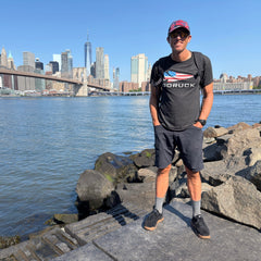 A man in shorts and sunglasses stands by the water with the NYC skyline and Brooklyn Bridge in the background, wearing a Cadre Hat - CORDURA®, crafted from rugged 500D CORDURA® and backed by the Scars Lifetime Guarantee for lasting adventure.