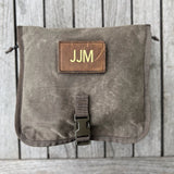 The Wire Dopp - Waxed Canvas pouch, featuring a leather "JJM" patch and buckle, is perfect for storing EDC essentials and is displayed on a wooden background.