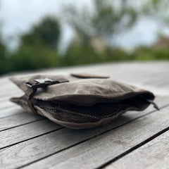 The Wire Dopp - Waxed Canvas, a compact brown pouch with a buckle, rests on a wooden table outdoors against blurred greenery—ideal for keeping your EDC organized.
