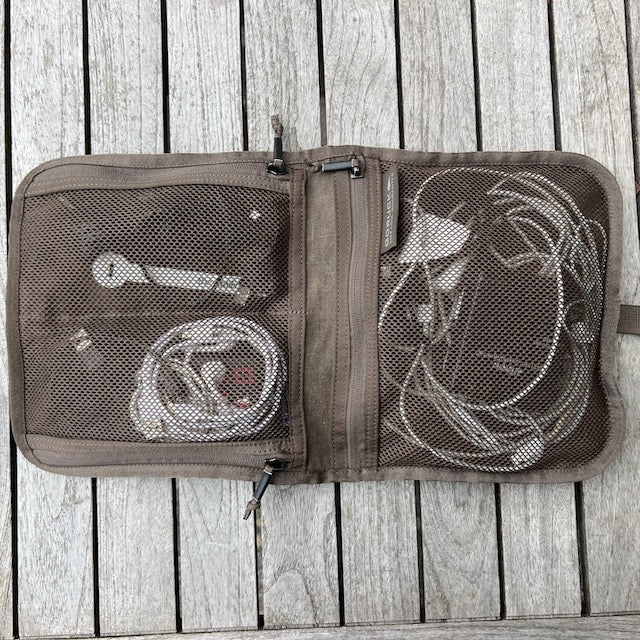 The Wire Dopp - Waxed Canvas is a brown mesh travel pouch open on a wooden surface, neatly holding white charging cables and adapters—perfect as an EDC travel cable organizer.