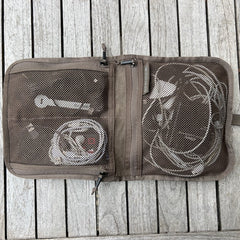 The Wire Dopp - Waxed Canvas is a brown mesh travel pouch open on a wooden surface, neatly holding white charging cables and adapters—perfect as an EDC travel cable organizer.