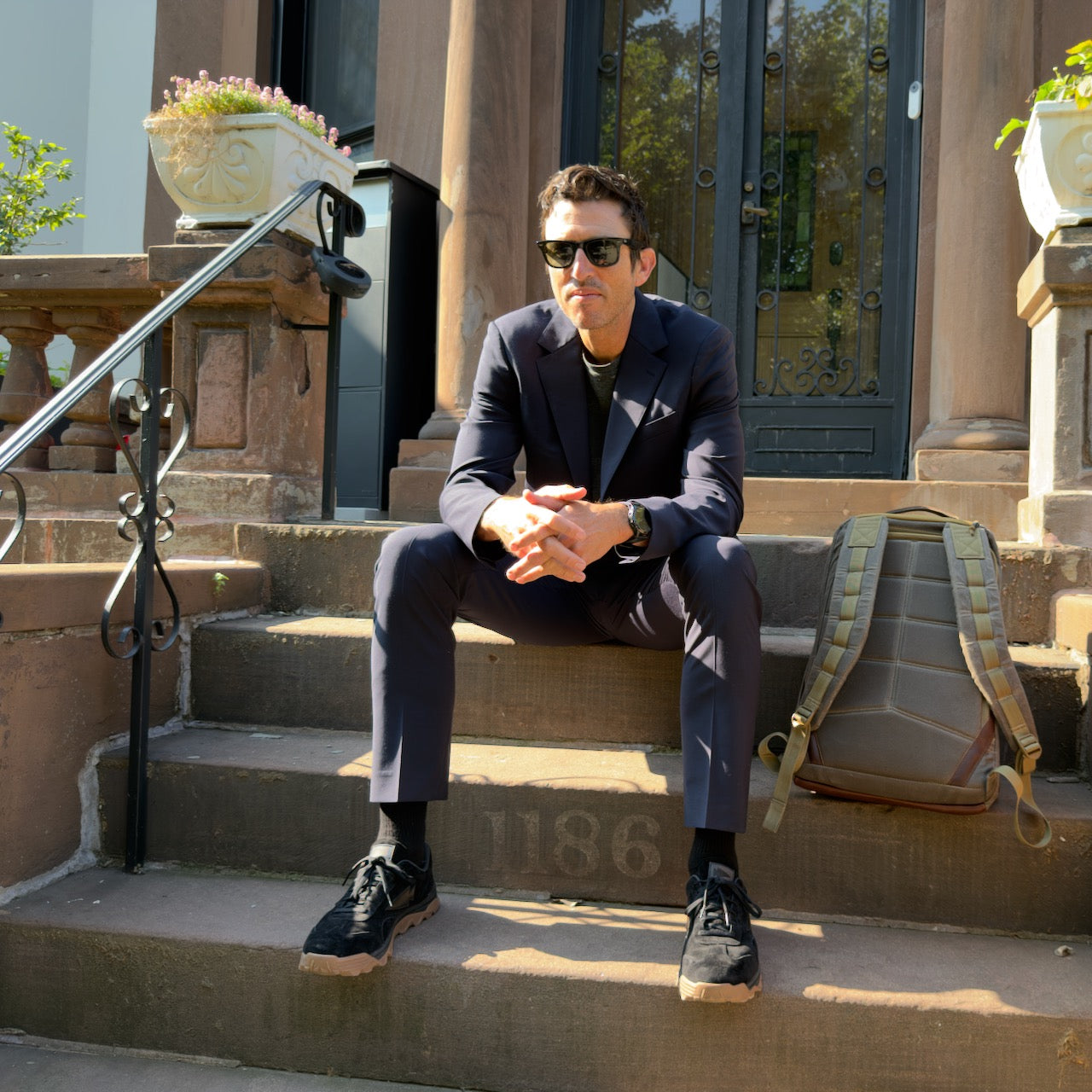 A man in a dark suit and sunglasses sits on brownstone steps beside the GR1 USA - Heritage Waxed Canvas rucksack, sunlight shining.