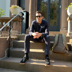 A man in a dark suit and sunglasses sits on brownstone steps beside the GR1 USA - Heritage Waxed Canvas rucksack, sunlight shining.