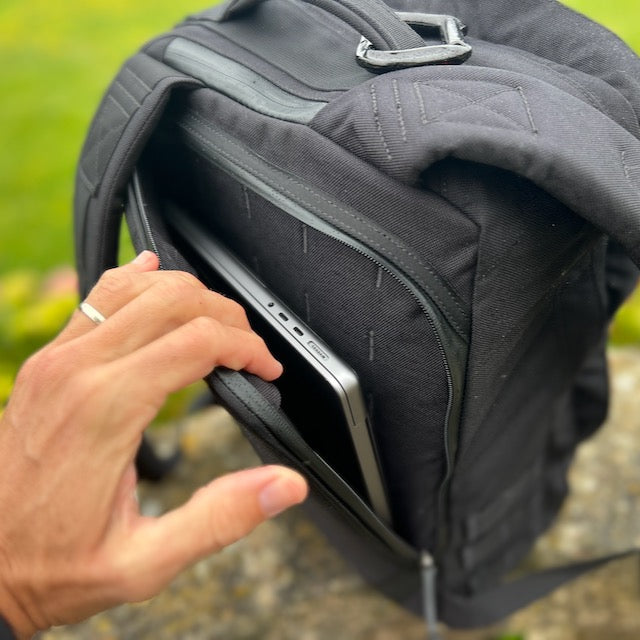 A hand slides a silver tablet into the front pocket of a black GR2 ruck outdoors—ideal for one bag travel and staying carry-on compliant.