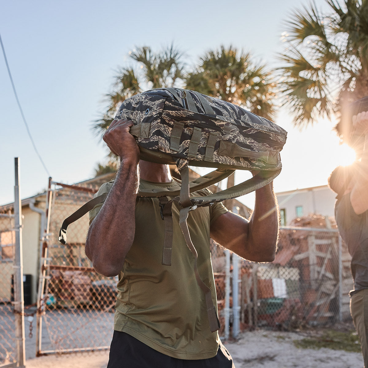 A person lifts the Rucker 4.0 backpack outdoors near a fence and palm trees, showcasing its rugged build and ruck plate compatibility.
