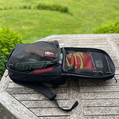 A GR2 ruck, opened on a wooden table outdoors amid greenery, features mesh compartments—ideal for one-bag travel and carry-on compliant adventures.