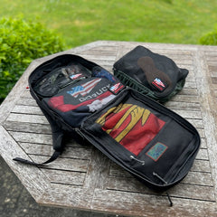 An open black GR2 backpack filled with clothes sits on a wooden table beside a packed pouch, ideal for one bag travel and carry-on compliant outdoor adventures.