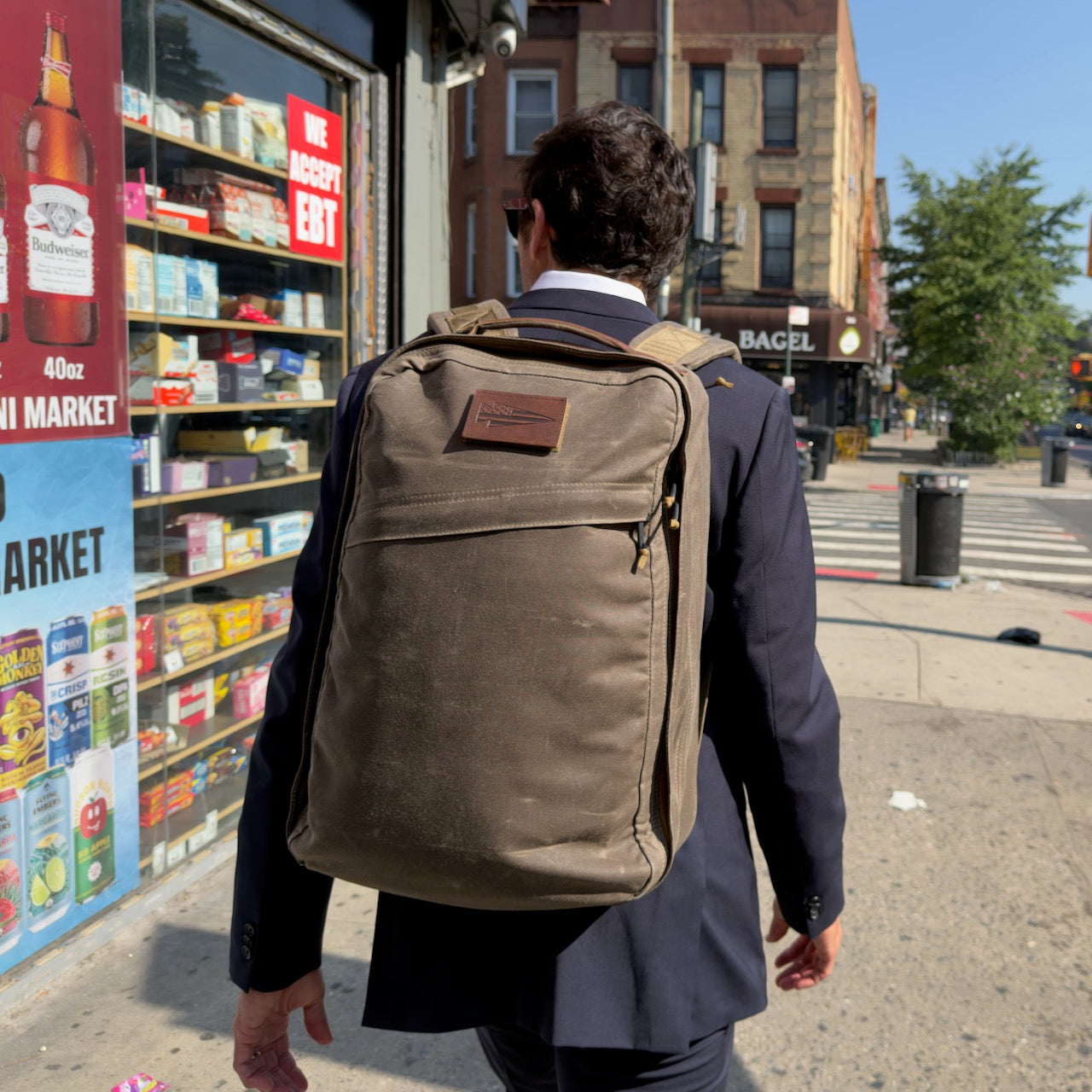 A man in a suit carrying the GR1 USA - Heritage Waxed Canvas backpack walks past a corner market on a sunny city street.