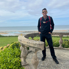 A man in black clothes with a red backpack and a Sternum Pouch - Cordura stands on a stone terrace overlooking the ocean and grassy area.