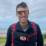 A smiling man by the coast wears sunglasses, a black jacket, and a red star-patterned backpack equipped with the Sternum Pouch - Cordura.