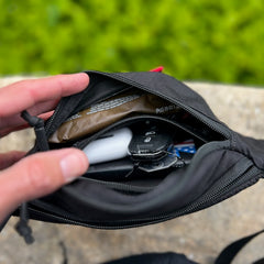 A hand opens the Tactical Fanny Pack - Cordura in black, revealing keys, a snack bar, AirPods, and other small essentials stored neatly inside.