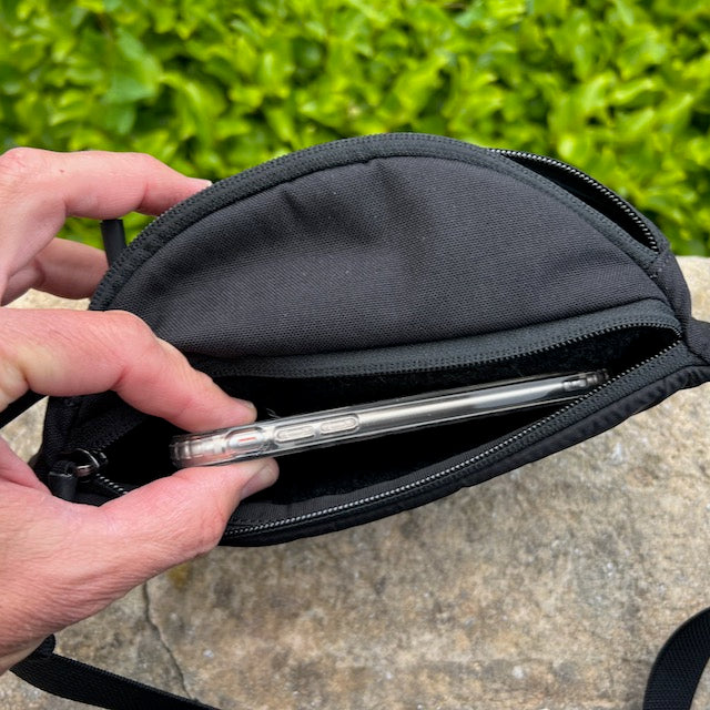 A hand places a smartphone into the padded pocket of the black Tactical Fanny Pack - Cordura, set on a stone surface with green foliage in the background.