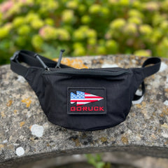 A black Tactical Fanny Pack - Cordura by GORUCK, featuring an American flag patch and a padded essentials pocket, rests on stone with greenery in the background.