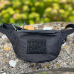 The Tactical Fanny Pack - Cordura, featuring a padded design and black arrowhead patch, rests on stone amid greenery—ideal for stylish, functional carry as a Glock 43 fanny pack.