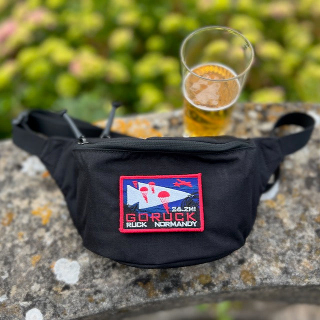A black Tactical Fanny Pack - Cordura with a colorful GORUCK Normandy patch is placed beside a glass of beer on a stone surface.