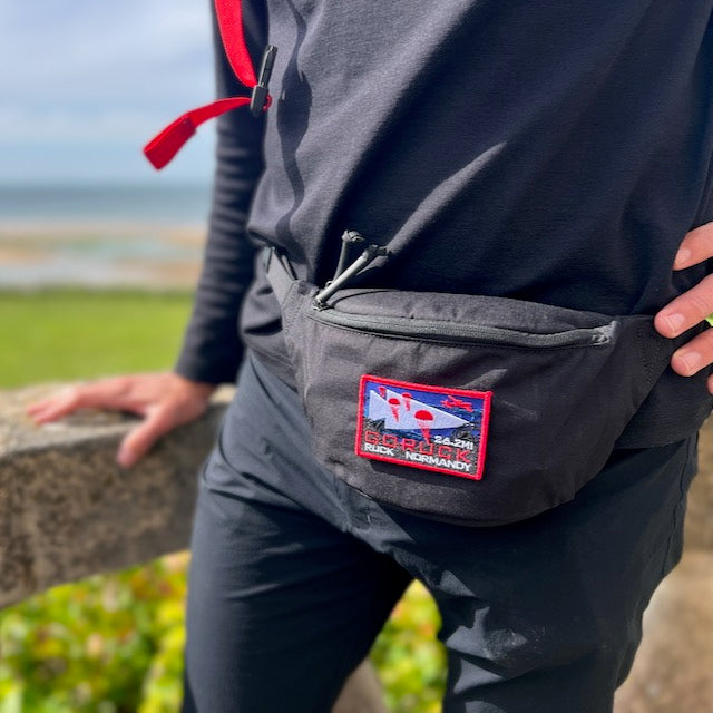 Person in black clothes wearing the Tactical Fanny Pack - Cordura, featuring a colorful patch, standing outdoors by a stone railing.