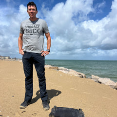 A man wearing Mackall 45 Black + Gum shoes and a gray “Embrace the Suck” shirt stands on a sandy beach with rocks and the ocean in the background.