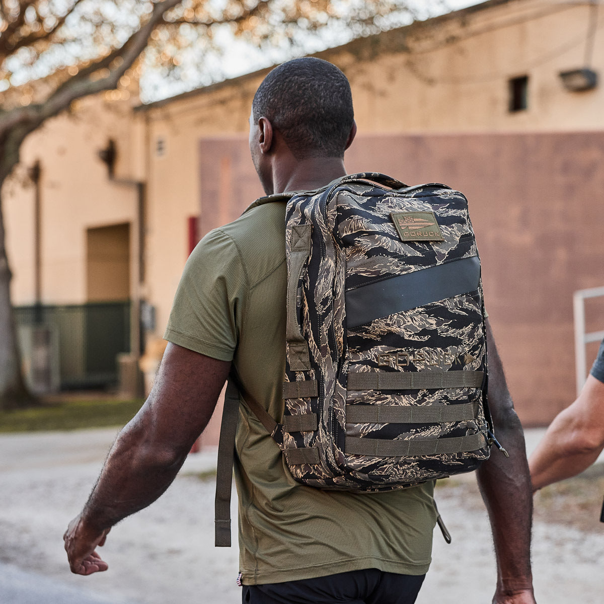A man wearing a green shirt and carrying the Rucker 4.0 backpack walks outdoors near a building and trees.
