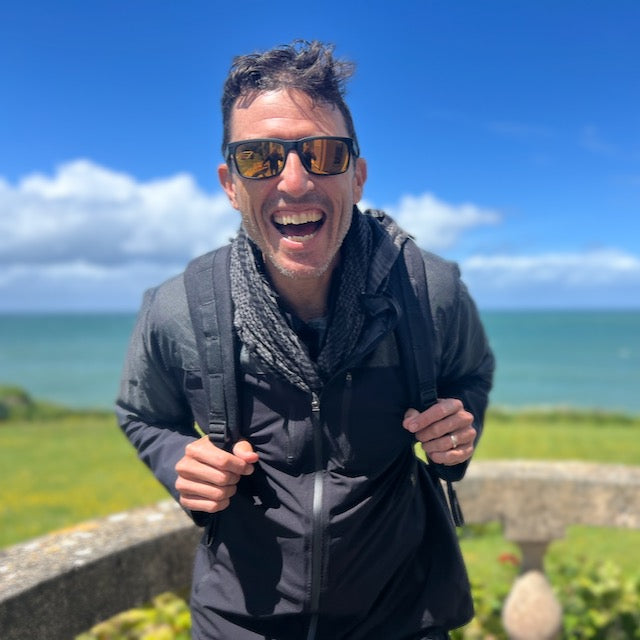 A man wearing sunglasses and a GR2 backpack enjoys one bag travel outdoors, with the ocean and blue sky behind him.