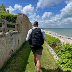 A person carrying the Rucker - Long Range walks a narrow seaside path under a partly cloudy sky, enjoying the benefits of distance rucking.