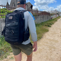 A man wearing a cap and backpack with a Simple Side Pocket walks on a dirt path near stone buildings and greenery under a partly cloudy sky.