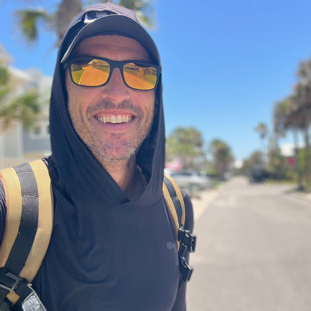 A man smiles outdoors in a Men's Sun Hoodie - ToughMesh, enjoying sunny weather by palm trees. The lightweight hoodie offers UPF 30+ sun protection, making it perfect for staying comfortable and stylish all day.