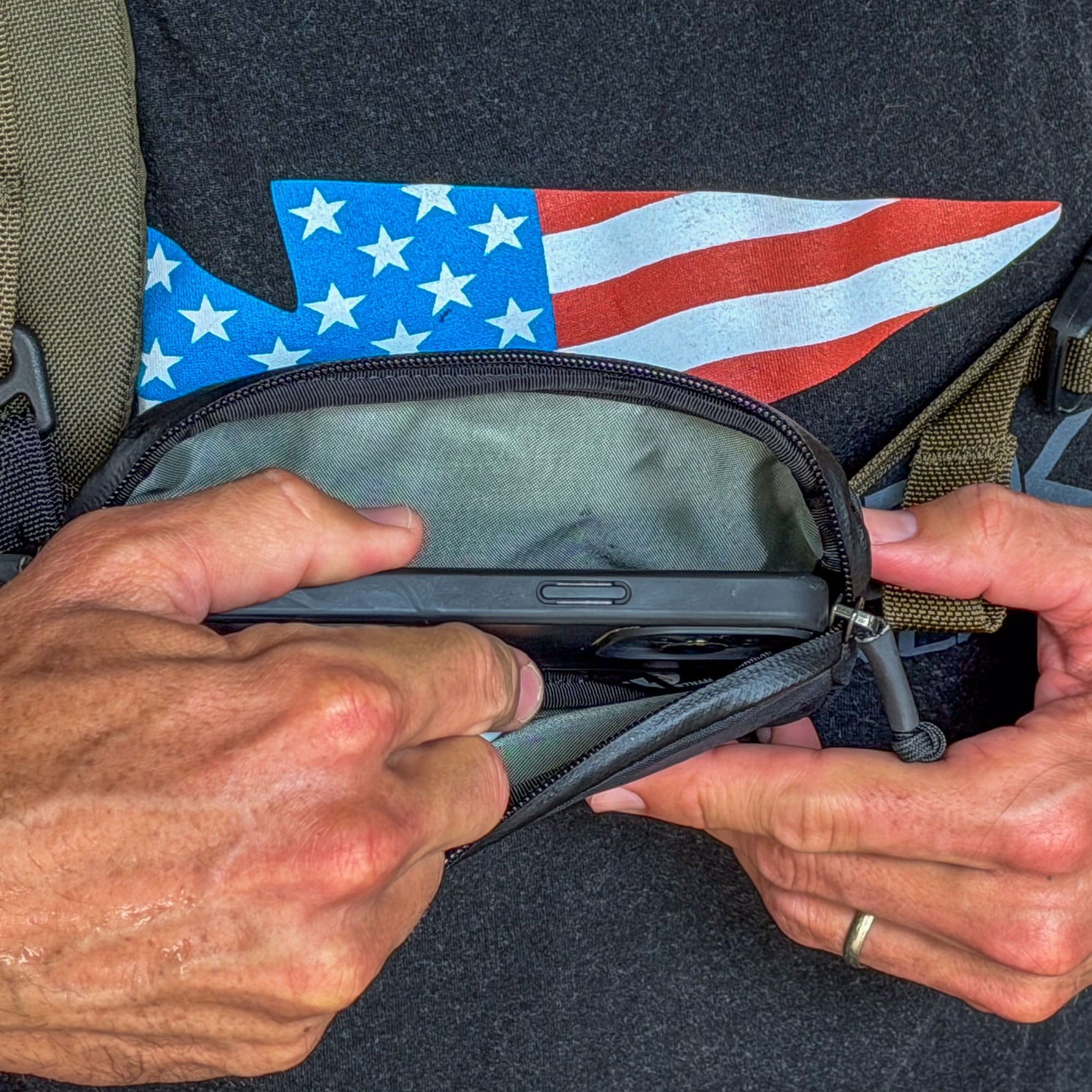 Hands placing a smartphone into the Sternum / Belt Pouch - Waterproof X-PAC for convenient access, with an American flag shirt in the background.