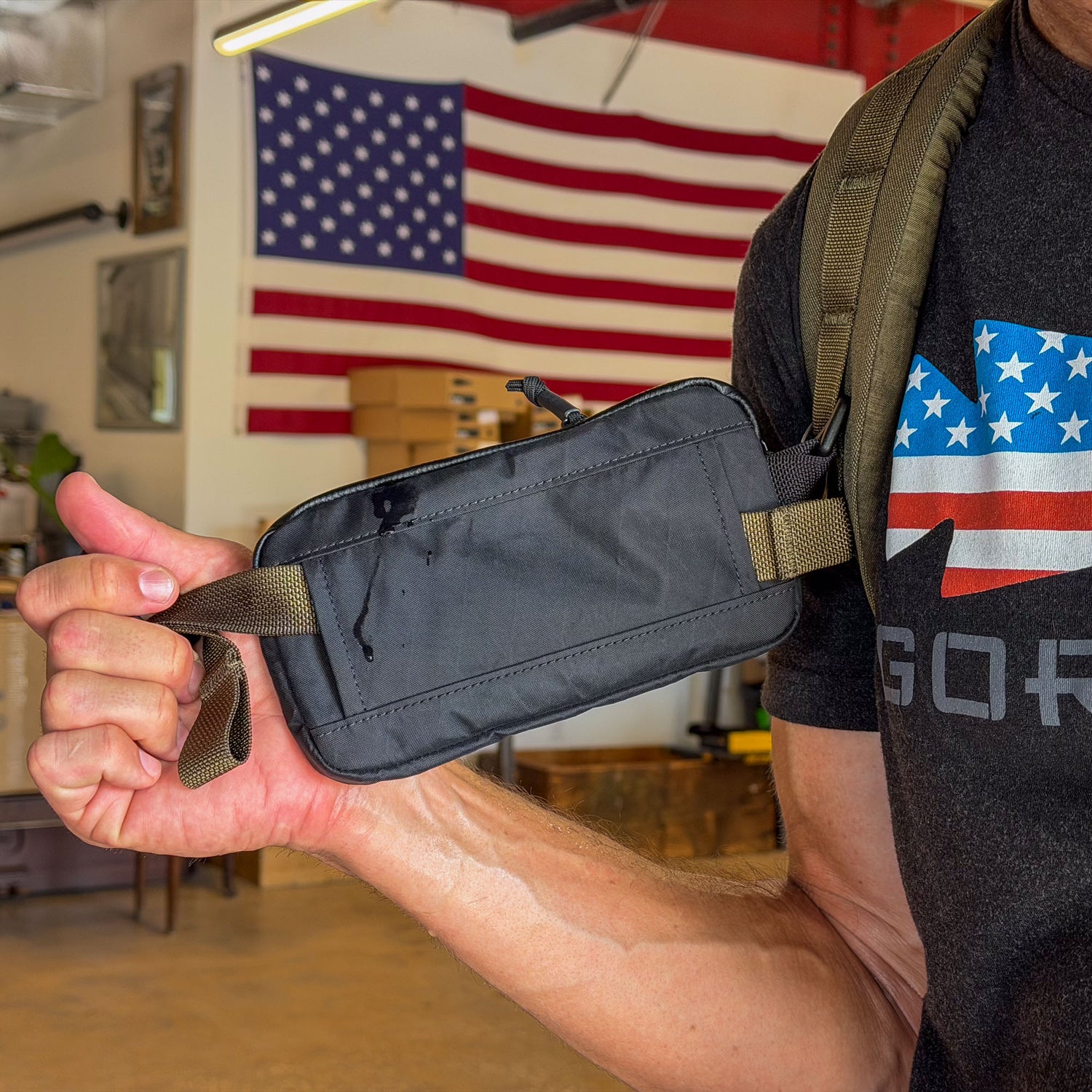 Someone holds the black Waterproof X-PAC Sternum / Belt Pouch, offering quick access; an American flag and a flag shirt appear in the background—ideal for rucking or outdoor adventures.