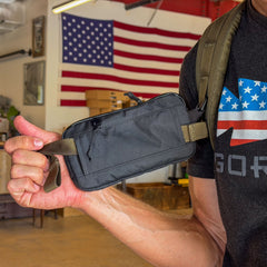 Someone holds the black Waterproof X-PAC Sternum / Belt Pouch, offering quick access; an American flag and a flag shirt appear in the background—ideal for rucking or outdoor adventures.