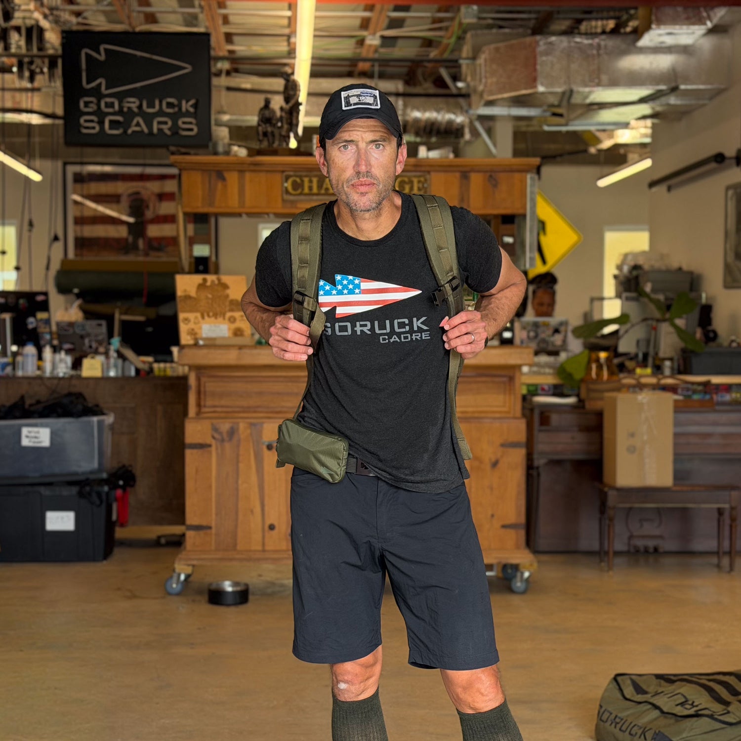 A man stands indoors in a workshop or gear store, wearing a GORUCK shirt and backpack with the Waterproof X-PAC Sternum / Belt Pouch, ready to test new rucking gear and hands-free storage solutions.