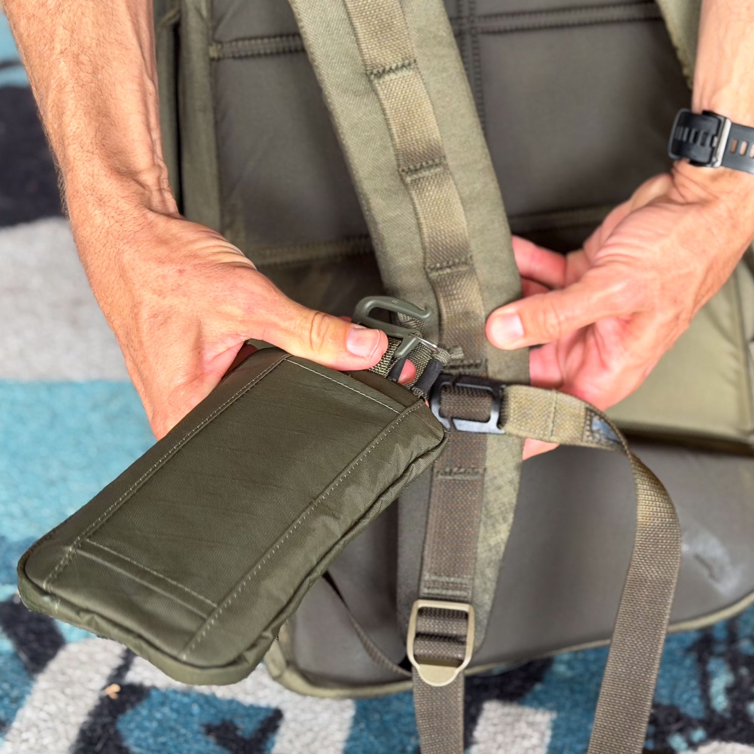 A person clips the olive green Sternum / Belt Pouch - Waterproof X-PAC onto a backpack strap—ideal for hands-free storage and organizing your rucking gear, with both hands visible.