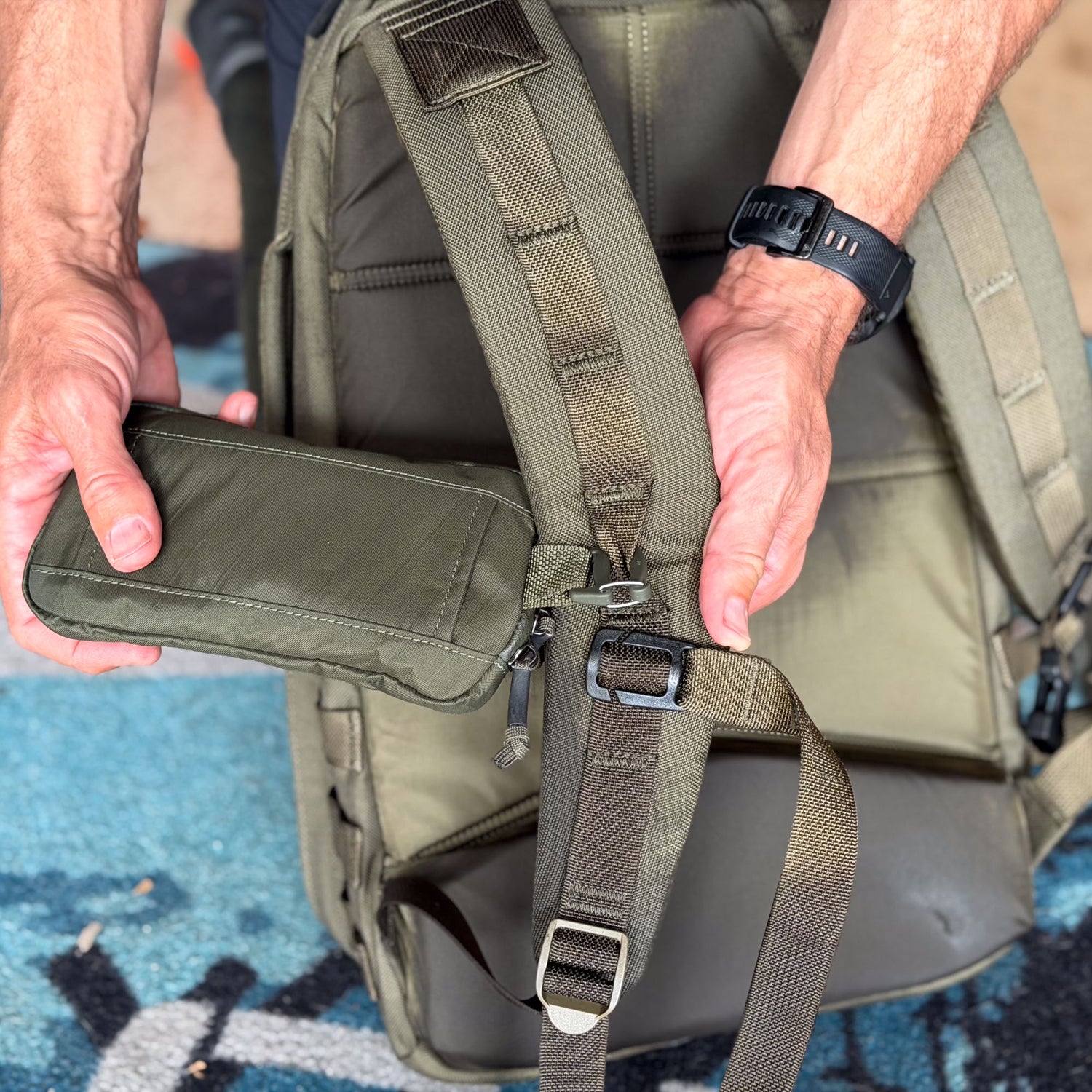 A person attaches the Sternum / Belt Pouch - Waterproof X-PAC in green to a strap on an olive backpack, adding hands-free, waterproof storage to their rucking gear.