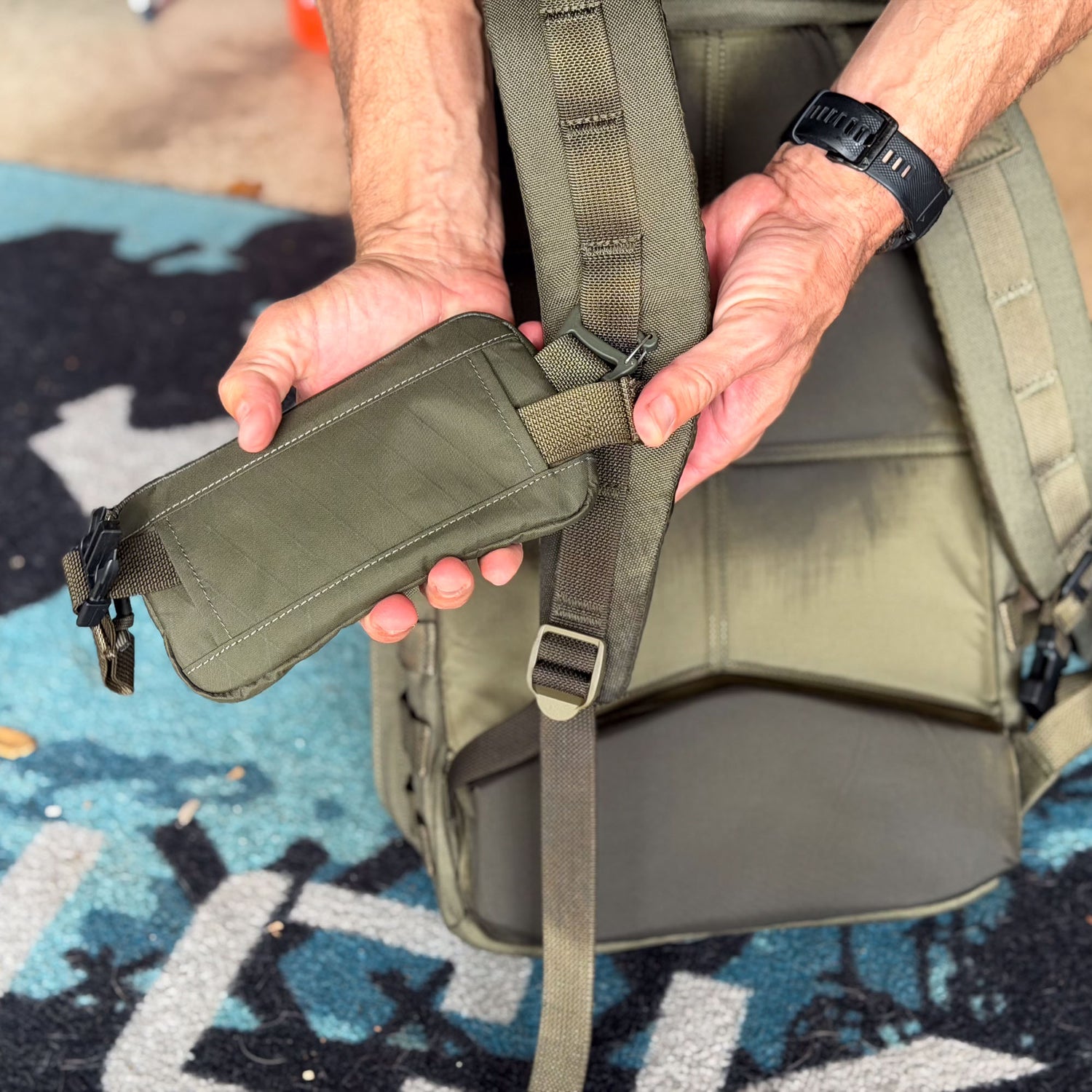Close-up of hands attaching the Sternum / Belt Pouch - Waterproof X-PAC in olive to a backpack strap outdoors, providing hands-free storage for essential rucking gear.