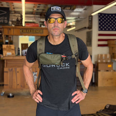 Wearing sunglasses and a cap, a man stands indoors with hands on hips, showcasing the Sternum / Belt Pouch - Waterproof X-PAC backpack; an American flag is visible in the background.