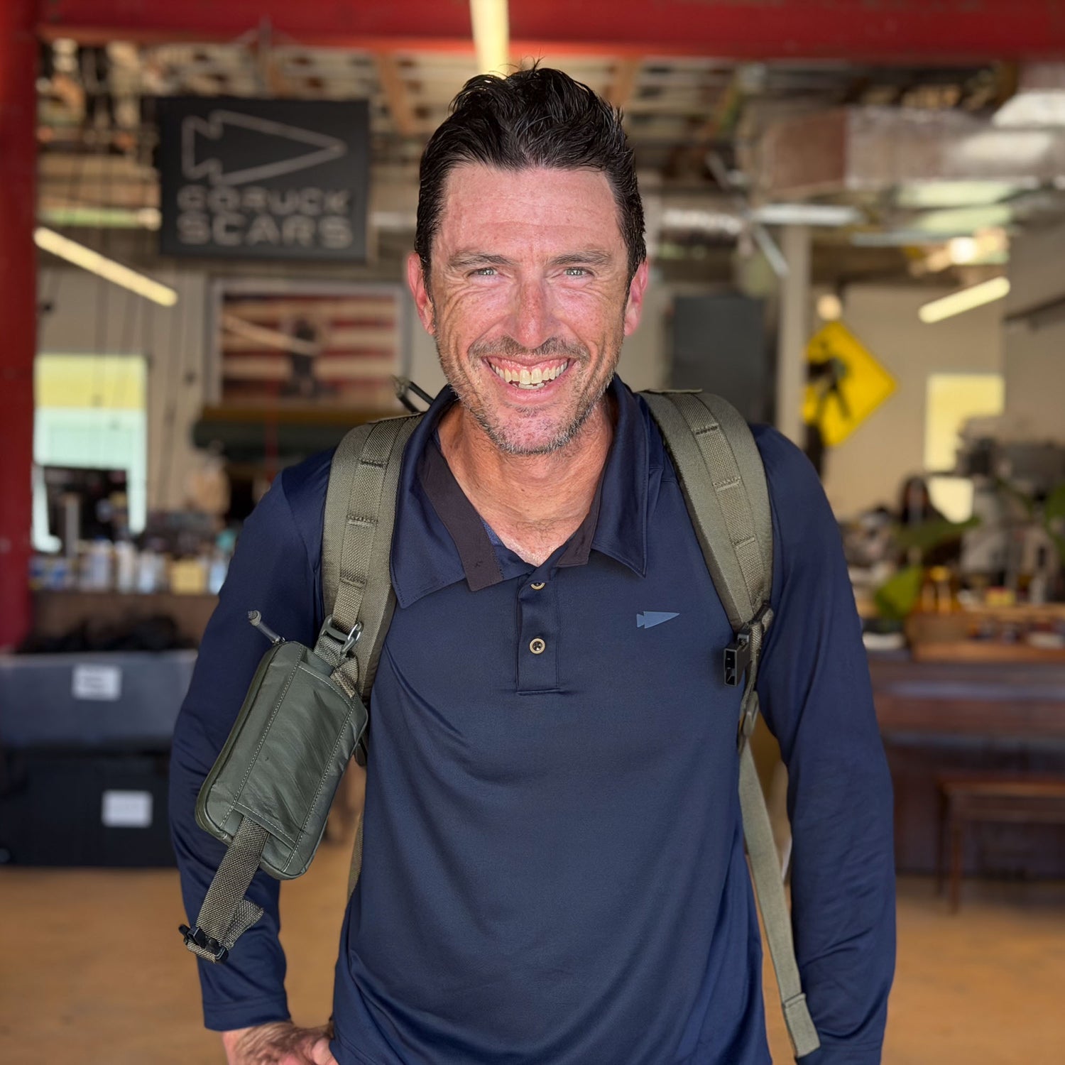 Man with backpack smiles in an indoor workshop, wearing the Men's Long Sleeve Rucking Polo in navy, made from EnduraKnit fabric, with red pillars and signs visible in the background.