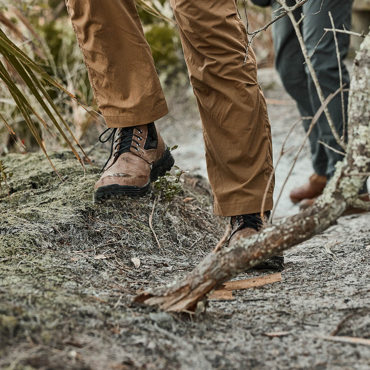 Close-up of people wearing MACV-2 Mid Top shoes with an aggressive triple compound outsole and brown pants, walking on a dirt trail surrounded by foliage and fallen branches.