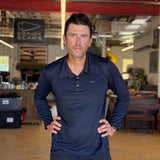 A man wearing the Men's Long Sleeve Rucking Polo, made in the USA, stands indoors with his hands on his hips, looking directly at the camera.