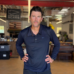 A man wearing the Men's Long Sleeve Rucking Polo, made in the USA, stands indoors with his hands on his hips, looking directly at the camera.