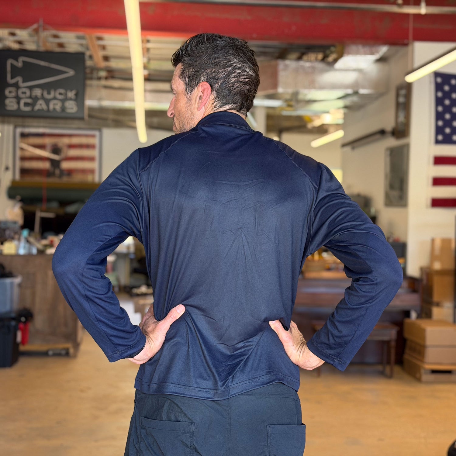 Man stands indoors in a workshop with hands on hips, facing away, wearing the Men's Long Sleeve Rucking Polo (navy), made in the USA.