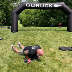 A man wearing Merino Challenge Socks - Ankle (No-Show) does push-ups on grass beneath a “GORUCK” inflatable arch at an outdoor event.
