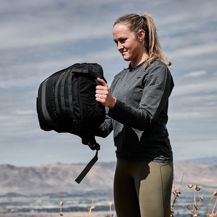 Amidst the stunning Italian Alps, silhouetted by a cloudy sky, a person outdoors carries a large black duffel bag while wearing the Men's Long Sleeve Performance Tee - ToughMesh.