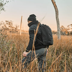 Wearing a black beanie and mask, someone in rugged Men’s Simple Pants - Midweight ToughDry® strolls through a grassy field at sunset with a backpack.