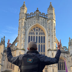 Wearing the Tac Hat - Heritage and a backpack, a person raises their hands skyward while facing a grand, ornate Gothic cathedral beneath a clear blue sky.
