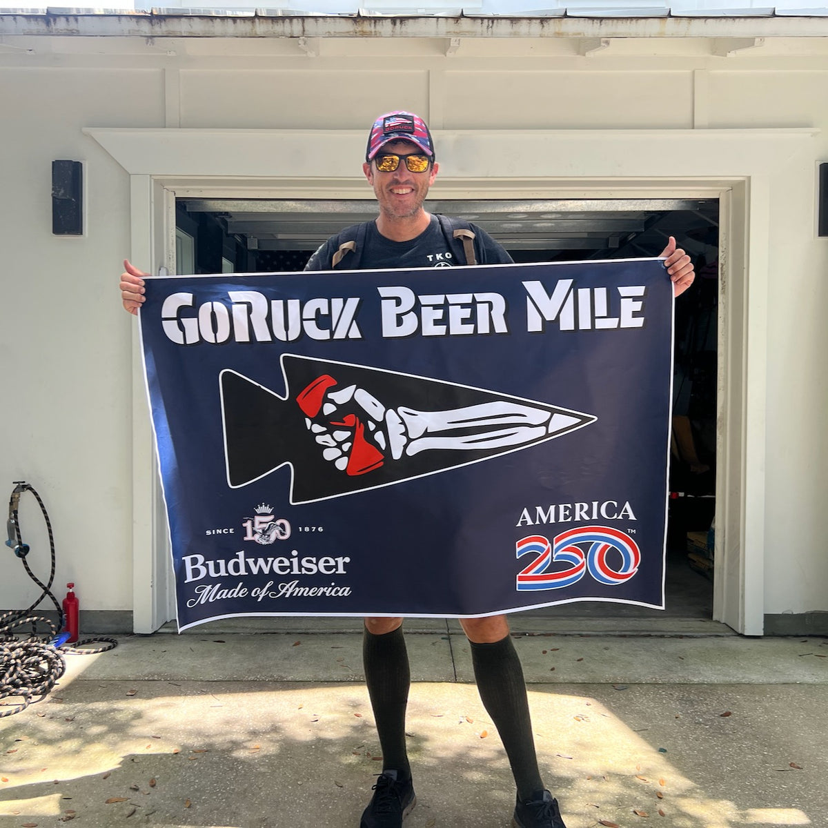 Smiling man wearing a Cadre Hat - CORDURA® and holding a “GoRuck Beer Mile” flag outside a garage on a sunny day.