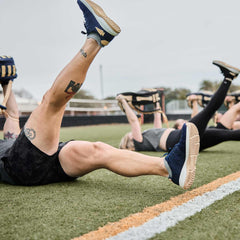 Group fitness session on grass field with people holding weighted bags and raising legs, wearing athletic shoes