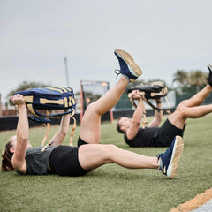 Two people doing leg raises on grass while holding blue and tan GORUCK backpacks in outdoor fitness training