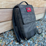 The Ruck Plate Carrier 3.0, featuring black camo with a red patch, is perfect for rucking workouts and is shown leaning against a wooden wall on gravel and leaves.